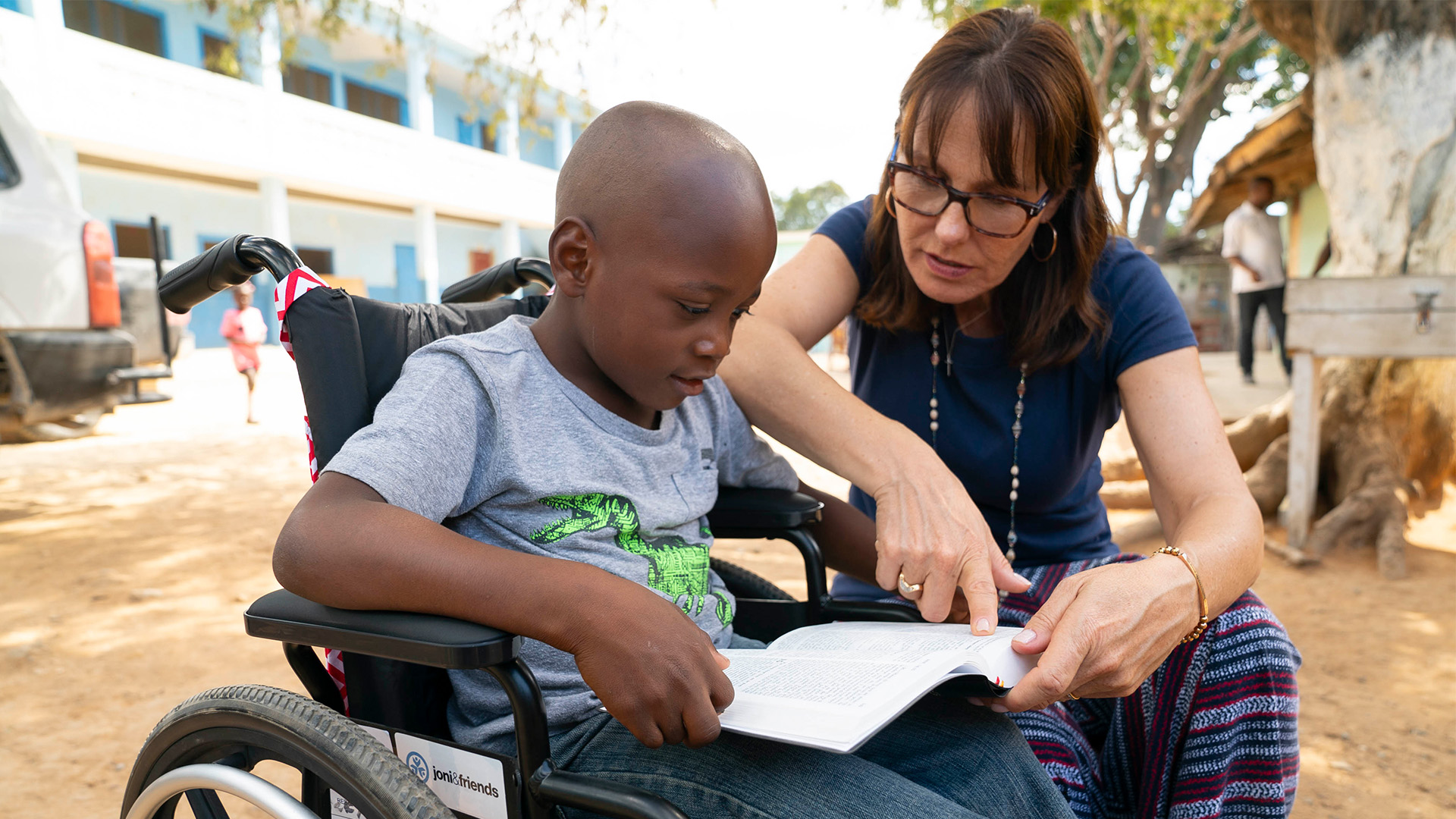 A boy in a wheelchair reads the Bible in his lap while a volunteer sits beside him, pointing to a scripture.
