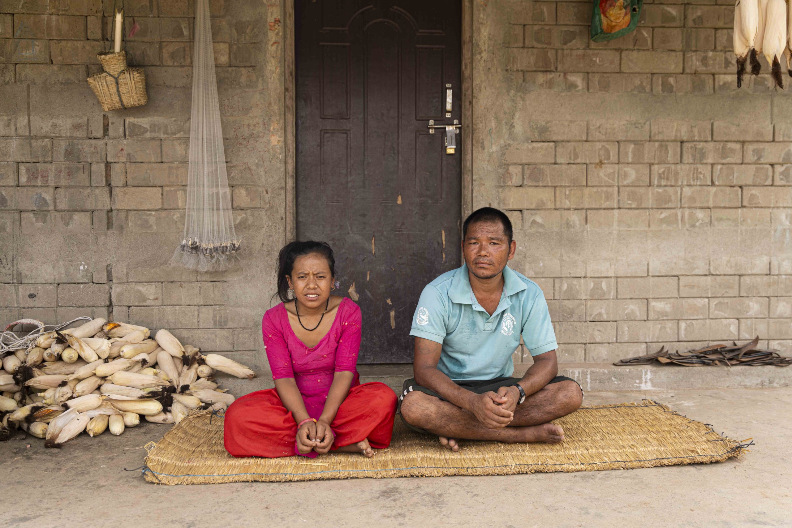 Padma and Biraj sitting next to each other on a straw mat, with ears of corn piled on their left side. Both of them are looking at the camera.