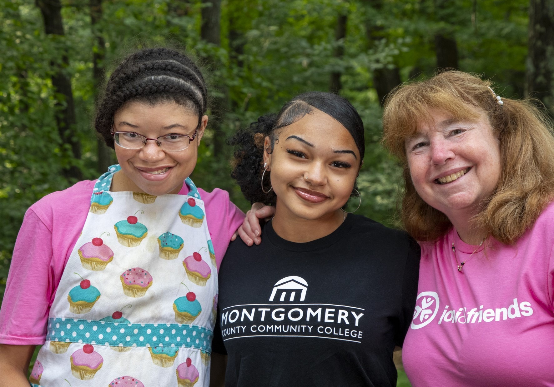 A picture of three women with their arms around each other and smiling at the camera. The one on the far left is a young girl wearing a cupcake apron, the girl in the middle is wearing a black college t-shirt and the woman on the right it wearing a pink Joni and Friends shirt.