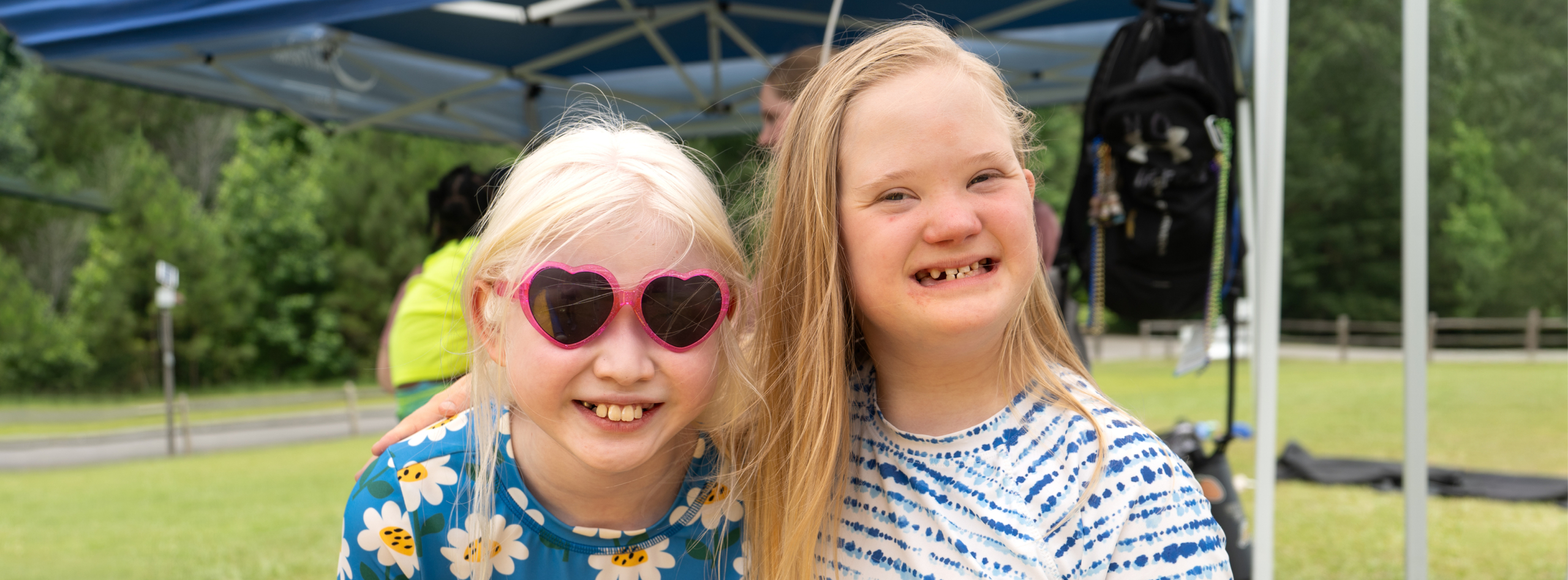 Two smiling girls outdoors at a family retreat—one wearing heart-shaped sunglasses and a floral shirt, the other in a tie-dye shirt.
