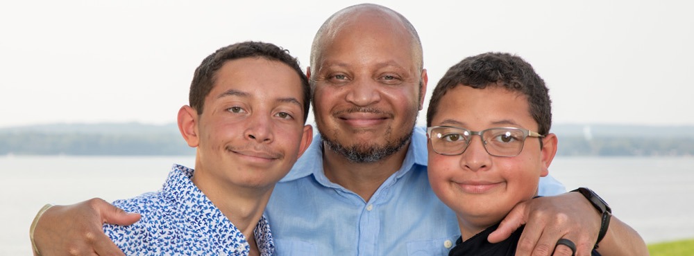 A father poses with his two boys in front of a lake.